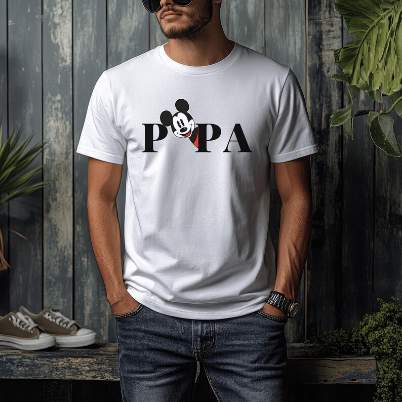 May include: White t-shirt with the word "PAPA" in black block letters. The "O" is replaced by a Mickey Mouse graphic in black, red, and white. The shirt is short-sleeved and made of a soft material.