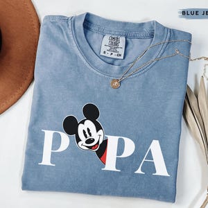 May include: A blue jean Comfort Colors t-shirt with a Mickey Mouse graphic. The graphic features Mickey's face and the word "PAPA" in white block letters. A gold necklace with a pendant is draped across the shirt.