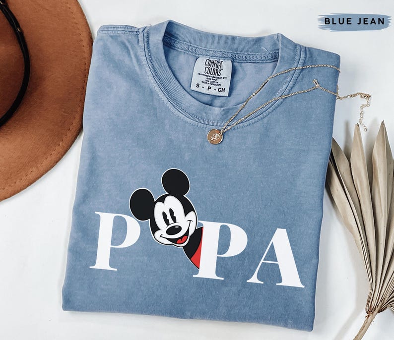 May include: A blue jean Comfort Colors t-shirt with a Mickey Mouse graphic. The graphic features Mickey's face and the word "PAPA" in white block letters. A gold necklace with a pendant is draped across the shirt.