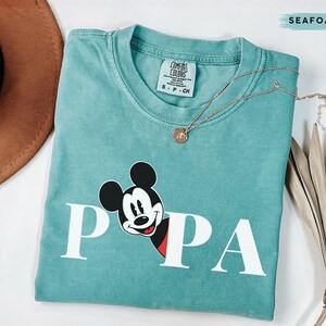 May include: Seafoam green t-shirt with the word "PAPA" in white block letters and a Mickey Mouse graphic. The shirt has a crew neck and short sleeves. A gold necklace with a pendant is draped across the shirt.