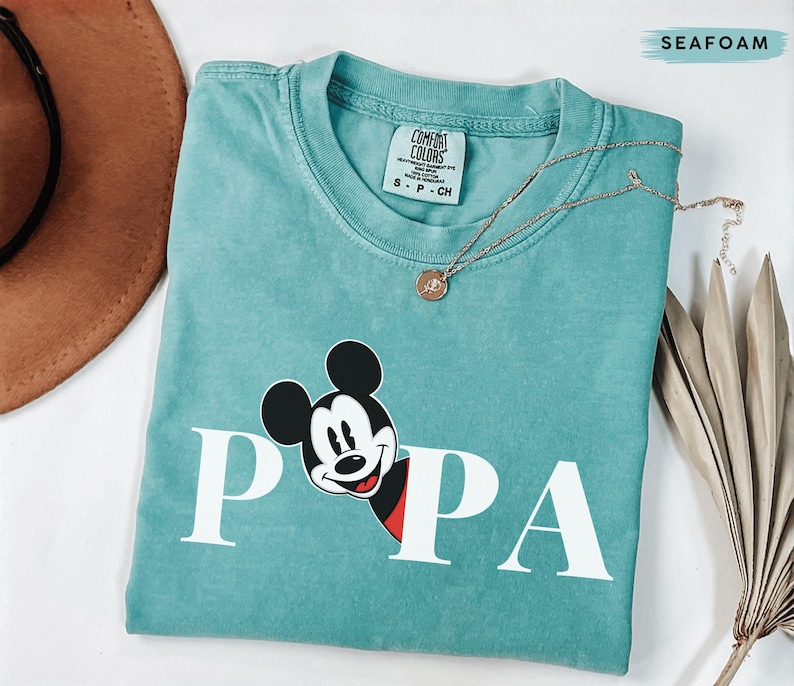 May include: Seafoam green t-shirt with the word "PAPA" in white block letters and a Mickey Mouse graphic. The shirt has a crew neck and short sleeves. A gold necklace with a pendant is draped across the shirt.