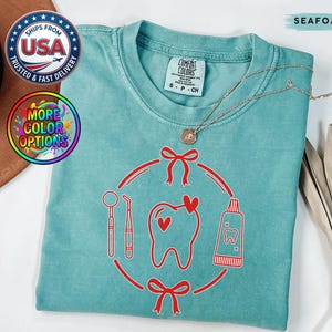 May include: Seafoam green t-shirt with a red dental-themed graphic. The graphic includes a tooth with a heart, dental tools, and a toothpaste tube. The shirt has a crew neck and short sleeves. The shirt has the text "COMFORT COLORS" on the tag.
