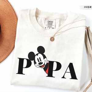 May include: Ivory-colored t-shirt with the word "PAPA" in black block letters. The "P" is partially replaced by a smiling Mickey Mouse graphic with a red and black outfit. A gold necklace is draped across the shirt.