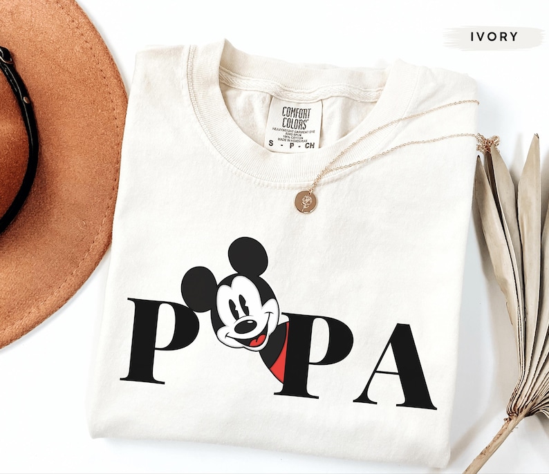 May include: Ivory-colored t-shirt with the word "PAPA" in black block letters. The "P" is partially replaced by a smiling Mickey Mouse graphic with a red and black outfit. A gold necklace is draped across the shirt.