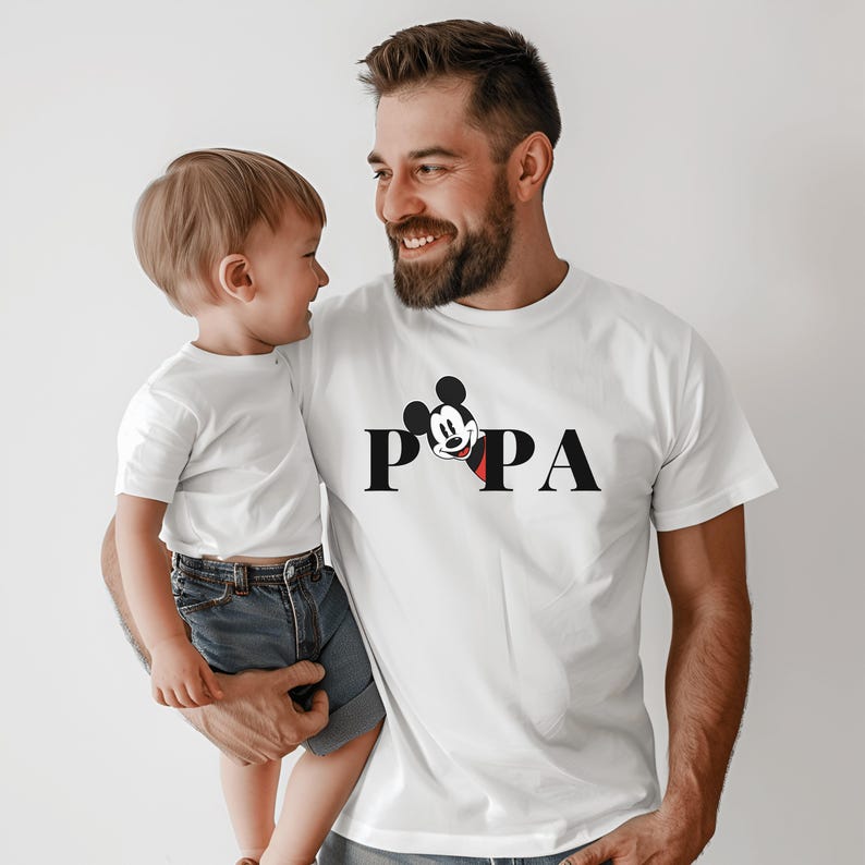 May include: White t-shirts with the word "PAPA" in black letters and a Mickey Mouse graphic. A man and a child are wearing the shirts. The child is wearing blue denim shorts.