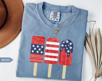 May include: A blue jean Comfort Colors t-shirt featuring three red, white, and blue popsicle designs. The popsicles have star and stripe patterns. A gold necklace is draped across the shirt.