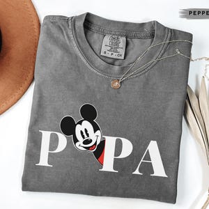 May include: A gray Comfort Colors t-shirt with the word "PAPA" in white block letters and a Mickey Mouse graphic. A gold necklace with a small pendant is draped across the shirt. A brown hat and dried plant are in the background.