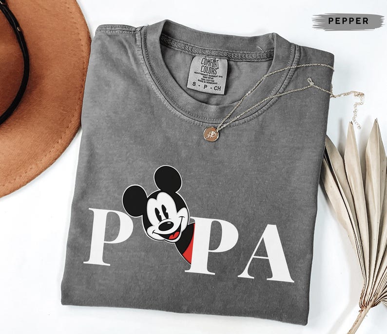 May include: A gray Comfort Colors t-shirt with the word "PAPA" in white block letters and a Mickey Mouse graphic. A gold necklace with a small pendant is draped across the shirt. A brown hat and dried plant are in the background.