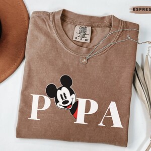 May include: Espresso-colored t-shirt with the word "PAPA" in white block letters and a Mickey Mouse graphic. The shirt has a crew neck and a Comfort Colors tag. A silver necklace with a small pendant is draped across the shirt.