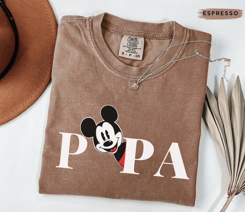 May include: Espresso-colored t-shirt with the word "PAPA" in white block letters and a Mickey Mouse graphic. The shirt has a crew neck and a Comfort Colors tag. A silver necklace with a small pendant is draped across the shirt.