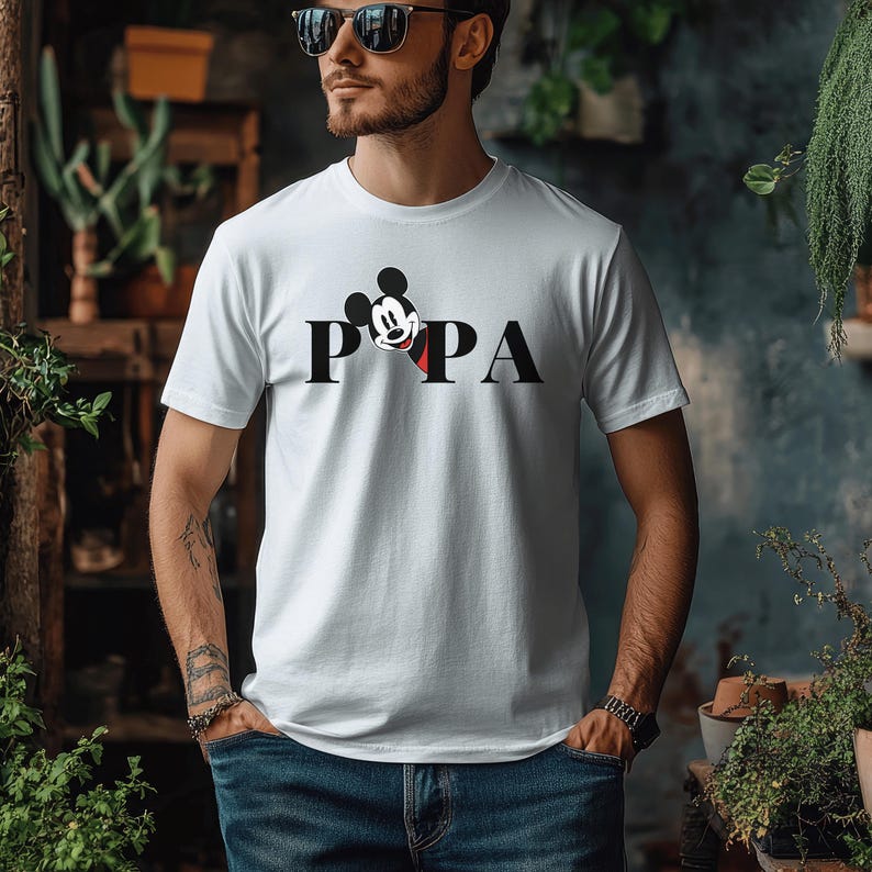 May include: White t-shirt with the word "PAPA" in black block letters. The "O" is replaced with a Mickey Mouse head. The shirt is short-sleeved and made of a soft material.