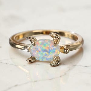 Op de afbeelding: Een goudkleurige ring met een ovaalvormige, iriserende opaalsteen in het midden. De ringband is ontworpen met vier kleine, getextureerde metalen uitsteeksels aan de zijkanten van de steen, die lijken op de ledematen van een zeeschildpad.
