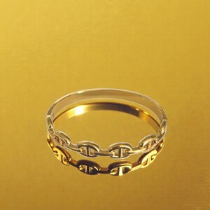 May include: A gold-toned bangle bracelet with a chain link design. The bracelet is reflected in a gold-colored surface. The chain links are oval-shaped and connected, creating a continuous loop. The bracelet is a piece of jewelry.