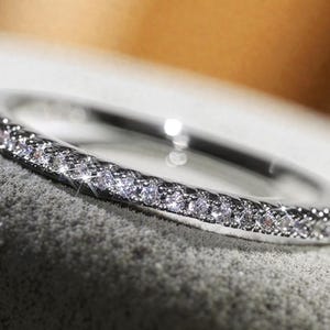 May include: A silver ring with a band encrusted with small, sparkling diamonds. The ring is set against a textured, light gray surface, with a soft, blurred background.