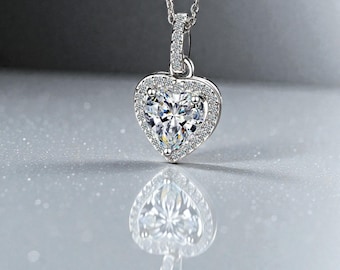 May include: A heart-shaped pendant necklace featuring a large, clear gemstone encircled by smaller, sparkling stones. The pendant is suspended from a fine silver chain. The necklace is presented on a reflective surface.