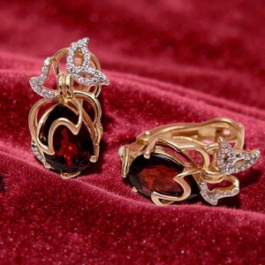Puede incluir: Un par de pendientes dorados con una piedra preciosa roja oscura en forma de lágrima. Los pendientes presentan un diseño de mariposa con pequeñas piedras transparentes. Las joyas se exhiben sobre una superficie de terciopelo rojo.