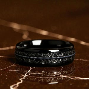 May include: A black ring with a detailed, patterned design around the centre. The ring has a glossy finish and sits on a brown surface with light-coloured veining. The ring's design is symmetrical and intricate.