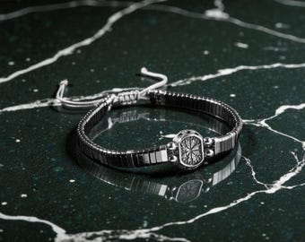 Könnte beinhalten: Ein silberfarbenes Armband mit rechteckigem Gliederdesign. Das Armband hat ein zentrales dekoratives Element mit einem komplizierten Muster. Der verstellbare Verschluss besteht aus weißer Kordel. Das Armband wird auf einer dunkelgrünen Oberfläche präsentiert.