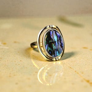 May include: A gold-coloured ring with an oval abalone shell displaying iridescent blues, greens, and purples. The ring has a double-band design around the stone, creating a unique and elegant look. The ring is set on a light-coloured surface.