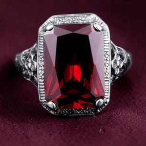 May include: A silver-toned ring featuring a large, rectangular, deep red faceted gemstone set in a frame with intricate engraved floral patterns along the band and a textured border surrounding the central stone, showcasing a polished finish.