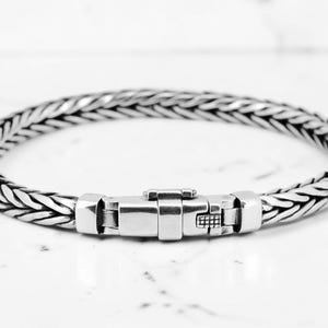 May include: A silver bracelet with a woven chain design. The bracelet features a secure clasp in the center. The jewelry is displayed on a white marble surface, creating a contrast with the metallic sheen.