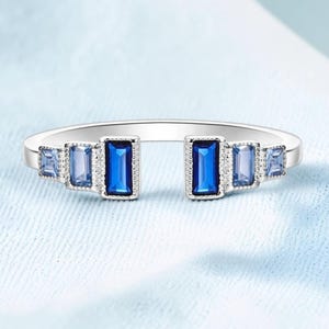 Può includere: Anello aperto in argento con una serie di pietre preziose blu. Il design presenta una grande pietra rettangolare blu scuro, pietre blu più chiare e minuscole pietre trasparenti. L'anello è aperto sul davanti.