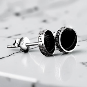 May include: A pair of silver stud earrings with a black circular center. The earrings have a decorative border and a post with a butterfly clasp. The earrings are on a white surface.