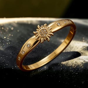 Puede incluir: Una pulsera dorada con un diseño de sol en el centro. La pulsera presenta pequeñas piedras preciosas transparentes y se encuentra sobre un fondo oscuro y texturizado. El sol está adornado con pequeñas piedras preciosas transparentes.