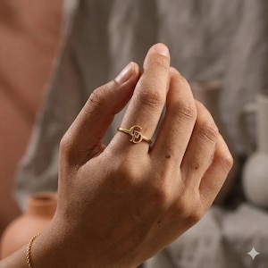 May include: A gold ring featuring the intertwined letters "G" and "P". The ring is worn on a finger, with a blurred background in neutral tones. The ring is likely made of metal.