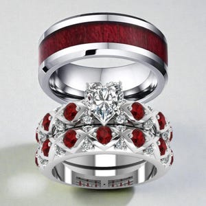 May include: A set of three rings. The top ring is silver with a wide, dark red wood inlay. The middle ring is silver with a heart-shaped diamond. The bottom ring is silver with red gemstones and small diamonds.