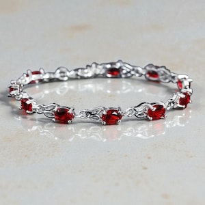 Può includere: Un bracciale in argento con pietre preziose ovali rosso scuro. Le maglie del bracciale presentano un delicato design floreale. Le pietre preziose sono uniformemente distanziate lungo il bracciale. Il bracciale è esposto su una superficie chiara.