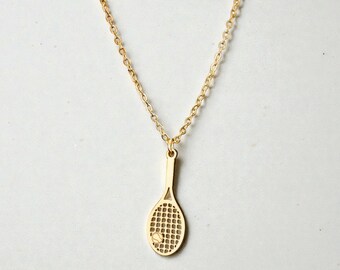 May include: Gold-coloured tennis racket pendant necklace. The pendant features a detailed racket design with a small tennis ball. The chain is delicate and the necklace is displayed against a white background.