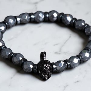 May include: A stretch bracelet featuring faceted metallic gray beads alternating with small smooth black spacers. A central black pendant shaped like a detailed helmet with small clear stones embedded along the crest hangs from the bracelet.