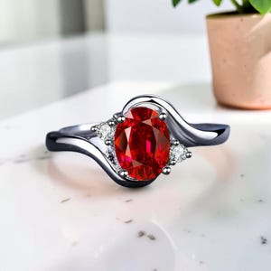 Könnte beinhalten: Ein silberner Ring mit einem großen, oval geschliffenen, leuchtend roten Edelstein als Mittelpunkt. Der Ring hat ein wirbelndes Design mit kleineren, klaren Akzentsteinen auf beiden Seiten des Hauptsteins. Der Ring liegt auf einer weißen Oberfläche.