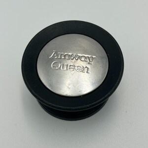 May include: A circular pot lid knob featuring a brushed silver metal center disc with the embossed text Amway Queen. The metal disc is surrounded by a thick, matte black circular rim, creating a functional handle component for cookware.