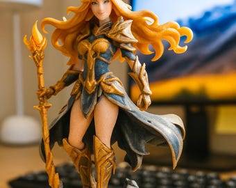May include: A detailed figurine of a female warrior with flowing blonde hair, wearing blue and gold armor, and holding a staff with a flame. The figure stands on a rocky base, showcasing intricate details and a dynamic pose.