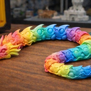 May include: A colorful, articulated dragon toy in a rainbow gradient. The dragon is made of plastic and has a detailed design with scales and a head. The toy is posed on a wooden surface.