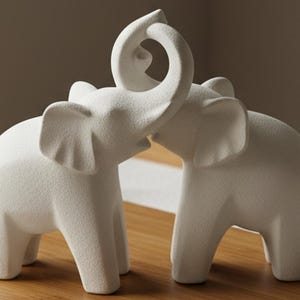 May include: Two white elephant figurines. One elephant has its trunk raised and intertwined with the trunk of the other elephant. The elephants are made of a textured material and are displayed on a wooden surface.