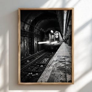 Puede incluir: Una impresión artística enmarcada en blanco y negro que muestra un tren de metro entrando en un túnel oscuro y arqueado. El tren tiene faros brillantes con desenfoque de movimiento, mientras que las vías están llenas de escombros.