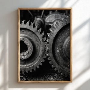 May include: A black and white framed wall art print featuring two large, weathered metal industrial gears interlocked. The gears show visible signs of wear, rust, and texture, with deep shadows between the teeth and a dark, gritty finish.