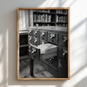 May include: A black and white framed print of a wooden library card catalogue cabinet with many drawers. One drawer is pulled open, revealing rows of white index cards. Metal label holders are attached to each drawer front with text labels.
