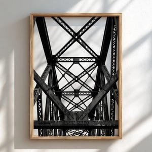 May include: A black and white photograph of a steel truss bridge structure, featuring a complex geometric pattern of intersecting beams and rivets, encased in a light-toned wooden frame that highlights the industrial architectural details.