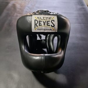 Op de afbeelding: Zwarte bokshoofdbescherming met het "CLETO REYES" logo in wit en goud. De hoofdbescherming is gemaakt van leer en heeft vulling rond het gezicht en de oren. Ontworpen om het hoofd te beschermen tijdens het boksen of andere vechtsporten.
