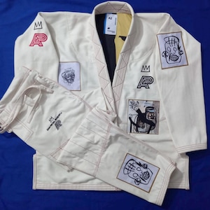 Puede incluir: Un gi de Jiu-Jitsu brasileño color hueso que incluye chaqueta y pantalón con costuras en contraste. El conjunto presenta varios parches bordados con diseños gráficos en negro y rojo, incluyendo coronas y el texto AP, con una etiqueta de talla A2.