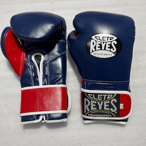 以下が含まれることがあります： 青と赤のCleto Reyesボクシンググローブ一組。白い縁取りが施されています。グローブの甲とリストストラップにはCleto Reyesのロゴがあり、リストラベルにはHecho en MexicoとProfessionalの文字が印刷されています。