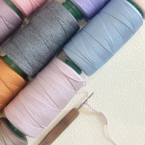 May include: An overhead shot of several spools of colorful thread in shades of pink, gray, blue, and orange. A crochet hook with a wooden handle and a partially completed chain stitch are also visible, suggesting a crafting project.