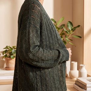 May include: A dark forest green open-front cardigan featuring a vertical ribbed knit pattern with alternating sheer, open-work panels. The garment has long sleeves with ribbed cuffs and a straight hemline, crafted from a lightweight, textured knit fabric.
