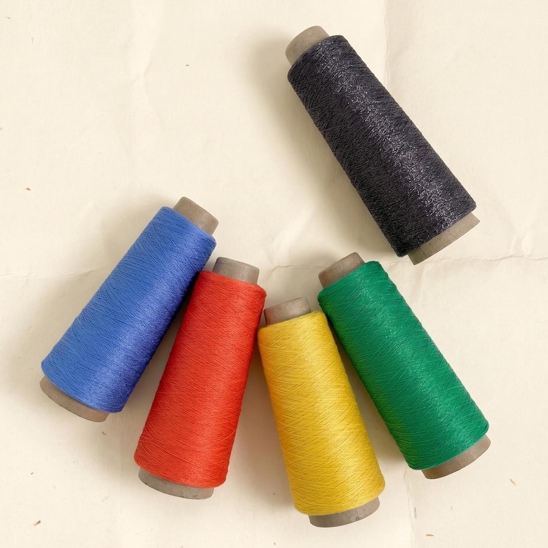 May include: Five conical spools of thread arranged in a fan shape. The colors include a dark charcoal grey, a vibrant blue, a bright red, a sunny yellow, and a rich green. Each spool features a central cardboard core and a textured thread finish.