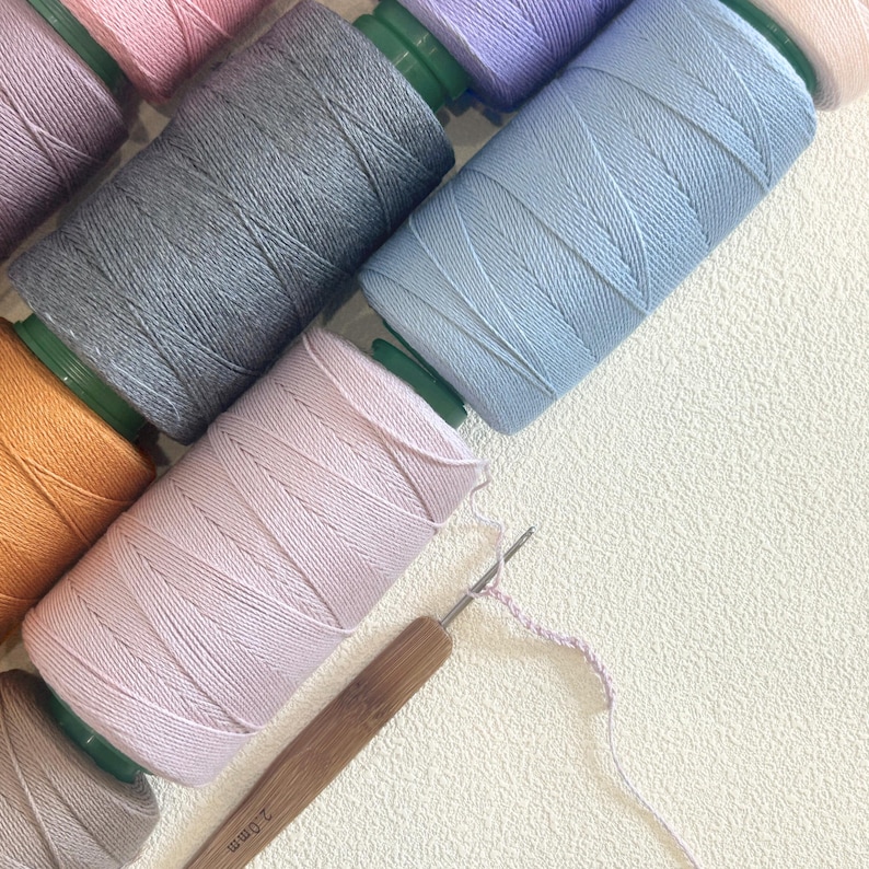 May include: An overhead shot of several spools of colorful thread in shades of pink, gray, blue, and orange. A crochet hook with a wooden handle and a partially completed chain stitch are also visible, suggesting a crafting project.