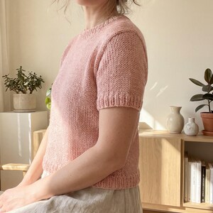 May include: A short-sleeved, crew-neck knit sweater in a soft, dusty pink color. The garment features a textured, hand-knit appearance with ribbed detailing along the neckline, sleeve cuffs, and the bottom hem, providing a structured fit.